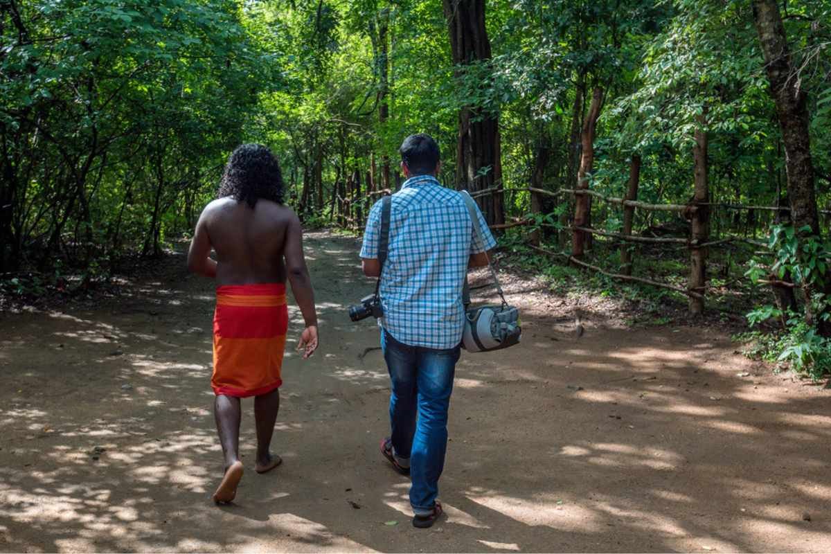 Un membre de la communauté Vedda décrit une tenue traditionnelle à un voyageur au milieu des forêts luxuriantes du Sri Lanka, symbolisant son lien profond avec la nature.